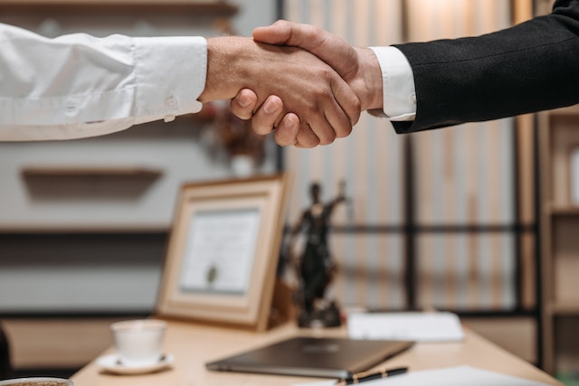 two people shaking hands across a desk with a status of lady justice in background